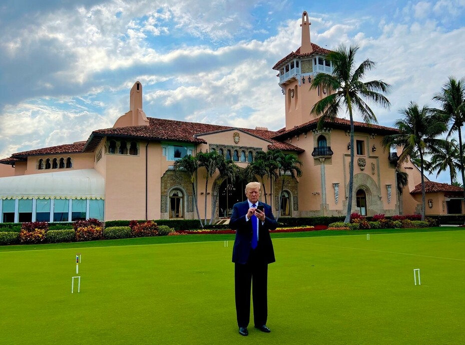 A man in a suit stands on a well-maintained lawn in front of a large, ornate building with a red-tile roof, palm trees, and a tower.
