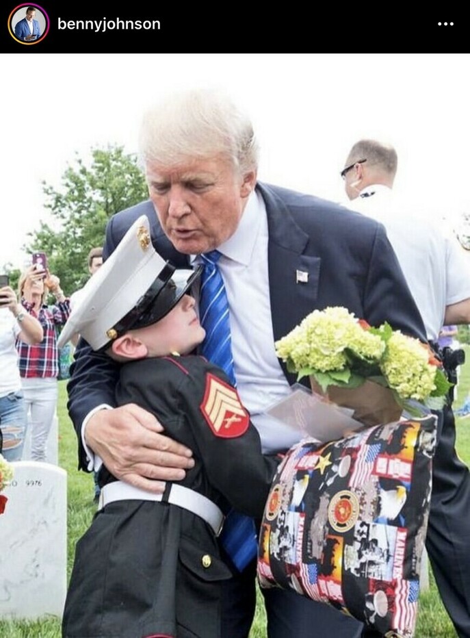 Former President Donald Trump hugging a young boy dressed in a Marine uniform, holding a bouquet of flowers and a patriotic pillow, at a cemetery with headstones in the background.