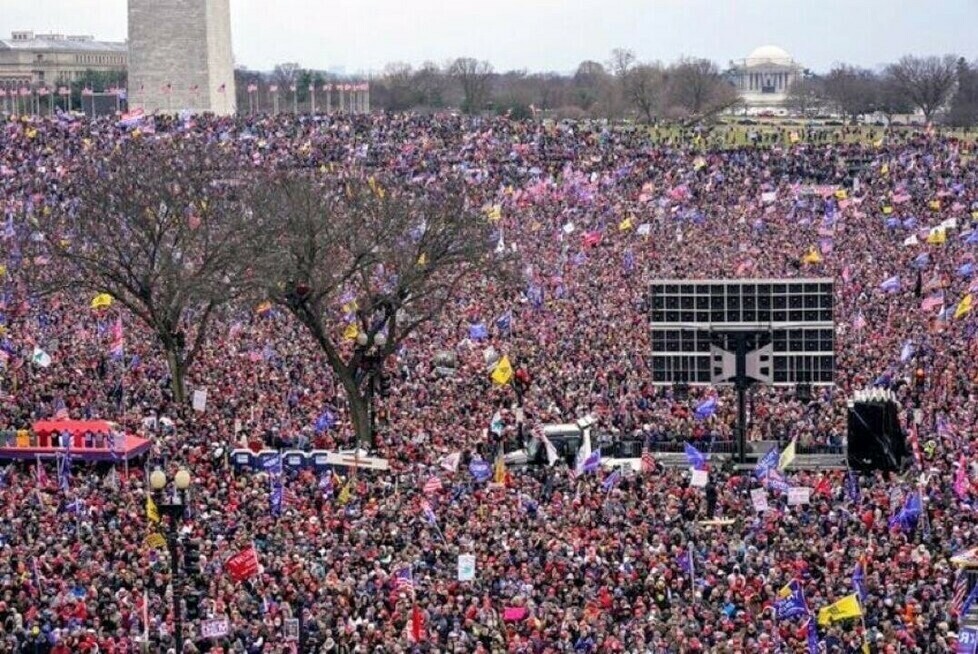 A densely packed crowd gathers near the Washington Monument, with numerous flags and signs visible among the people. The Jefferson Memorial is visible in the background, and some trees and a large screen are seen in the mid-ground. The gathering appears to be taking place in a large outdoor area with a mix of colorful clothing worn by the crowd.