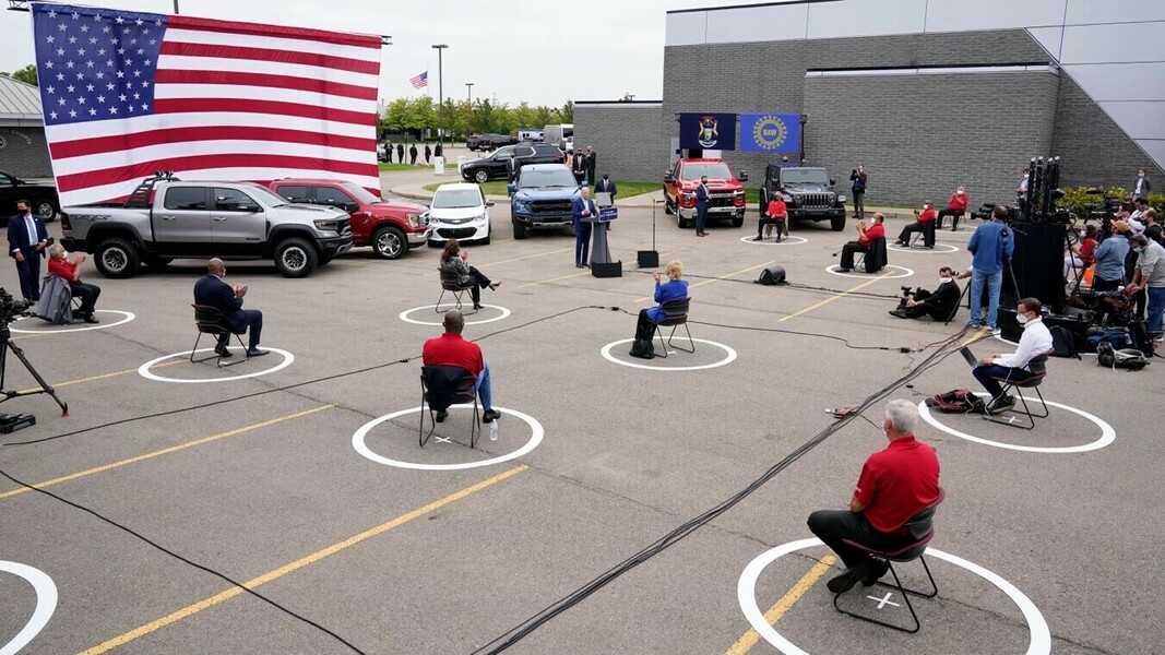Outdoor event with participants practicing social distancing guidelines, seated in chairs within white circles. Large American flag backdrop and several cars parked behind the speaker's podium. Stage area includes a person speaking at the podium and attendees wearing masks, along with a filming crew capturing the event. Banners of the state of Michigan and the UAW (United Auto Workers) are displayed on the building wall.