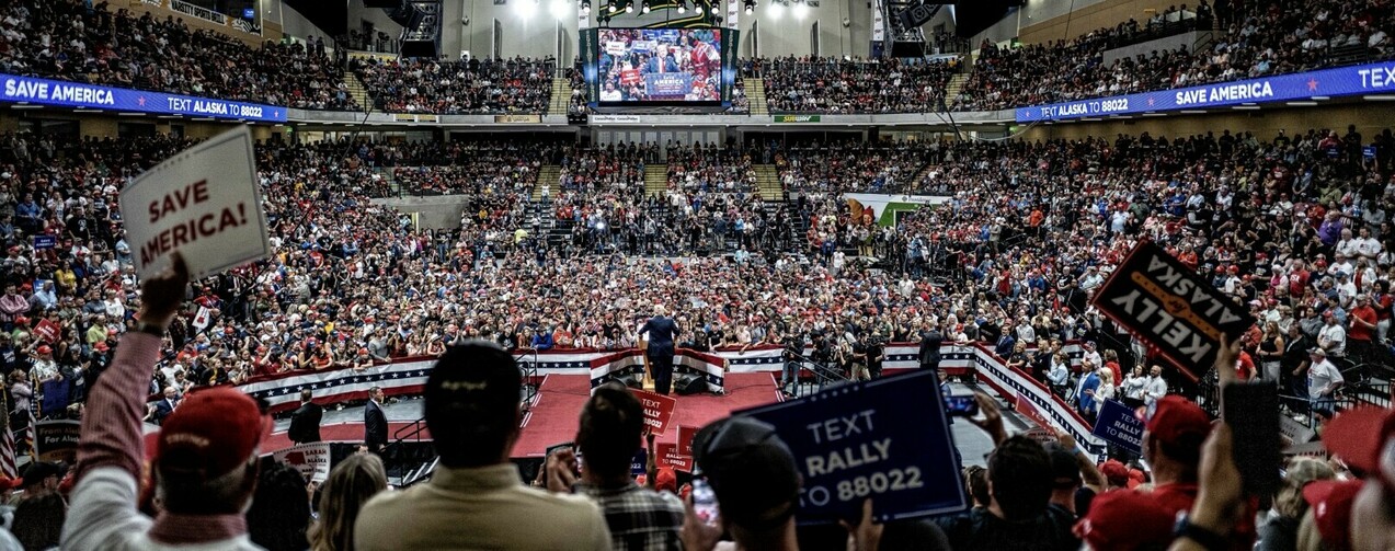 A large crowd gathers in an indoor arena for a political rally. Several people hold up signs, including one reading "SAVE AMERICA" and another "KELLY FOR ALASKA." There is a stage decorated with American flags, and a speaker is addressing the audience. Large screens display the event, and banners with messages such as "TEXT ALASKA TO 88022" are prominently displayed. The arena seats are filled with spectators.