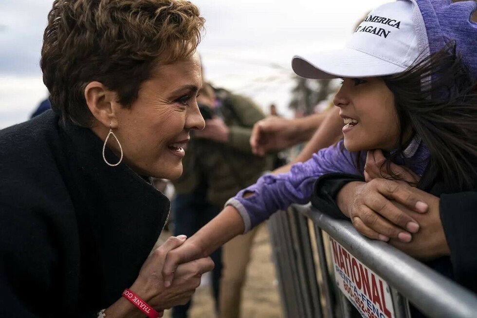Woman with short hair and teardrop earrings smiling and shaking hands with a young girl wearing a "Make America Great Again" cap and purple hoodie while standing by a metal barrier.
