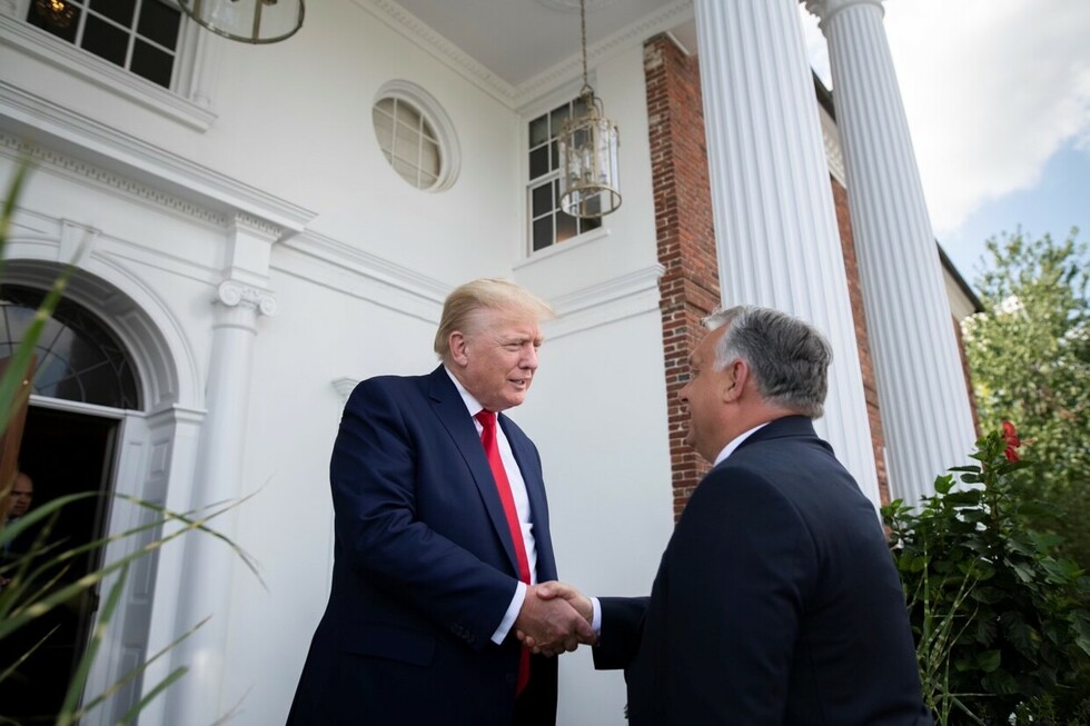Two men in suits shake hands outside a white building with columns and a brick exterior. One man has blonde hair and wears a dark blue suit with a red tie, while the other has grey hair and wears a dark suit. A glass lantern hangs above them and green plants are visible in the foreground.
