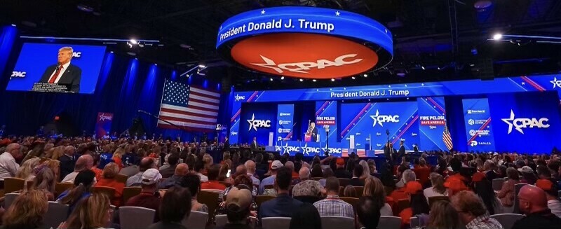 Former President Donald J. Trump speaking at the Conservative Political Action Conference (CPAC) event, with a large crowd in attendance, an American flag displayed prominently, and multiple screens projecting his image and the CPAC logo in the background.