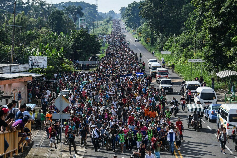 Large crowd of people walking along a road, surrounded by dense foliage and passing several vehicles. The image depicts a long, orderly procession stretching into the distance, with some bystanders observing from the sides.