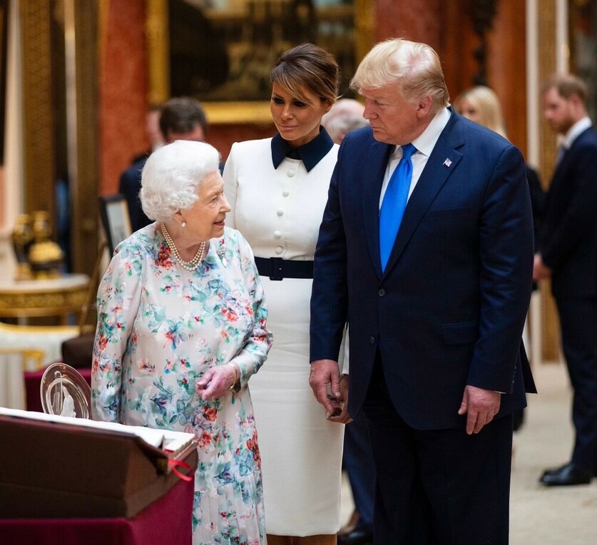 Three people conversing in an ornate room, with a woman in a floral dress smiling, another in a white dress with black details watching attentively, and a man in a blue suit with a blue tie looking on. A framed portrait and various decorative items are visible in the background.