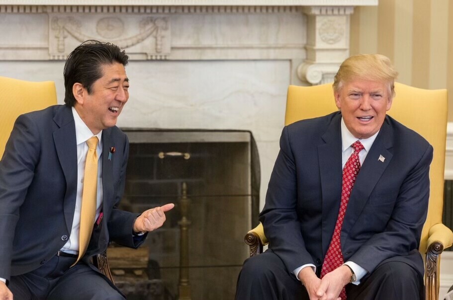 Two men in suits sitting in yellow chairs, smiling and engaged in conversation, with a fireplace in the background.