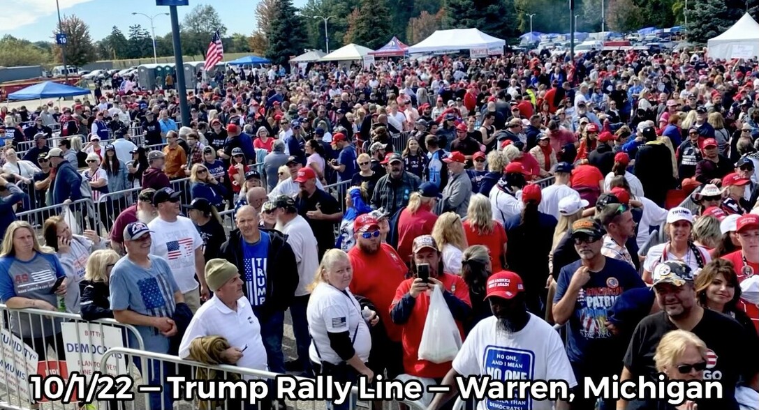 Crowd of people gathered at a Trump rally in Warren, Michigan on October 1, 2022, wearing various "Trump" apparel and holding signs; tents and American flags in the background.