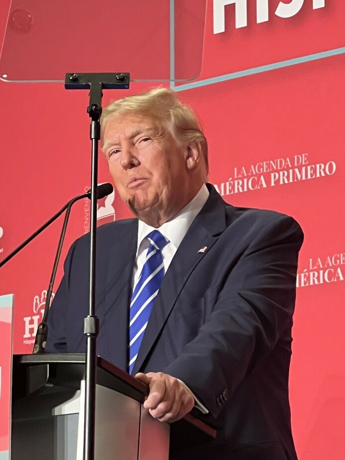 A man in a dark suit and blue striped tie speaks at a podium against a red backdrop displaying the text "LA AGENDA DE AMÉRICA PRIMERO" and "HISPANIC."