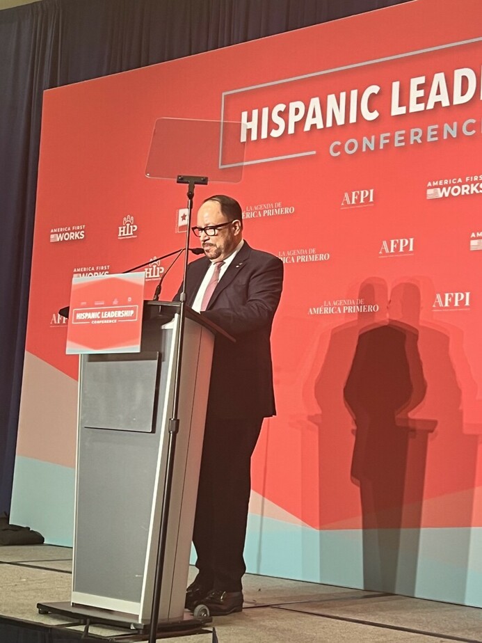 A man speaks at a podium during the Hispanic Leadership Conference, standing in front of a red backdrop with logos including America First Works and AFPI.