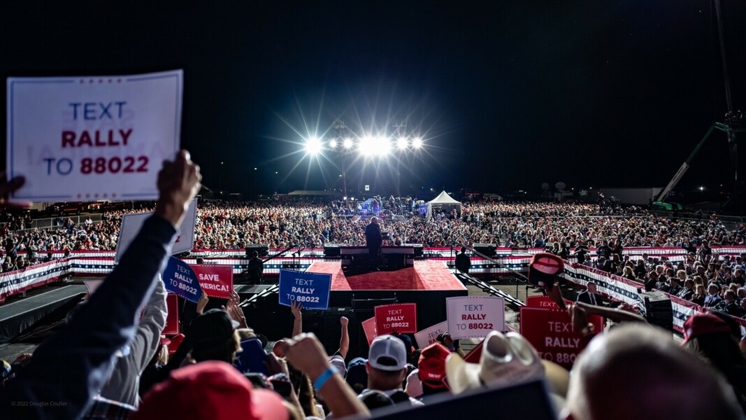 Crowd at a nighttime political rally with signs saying "Text RALLY to 88022" and "Save America" and a speaker on a stage with bright lights in the background.