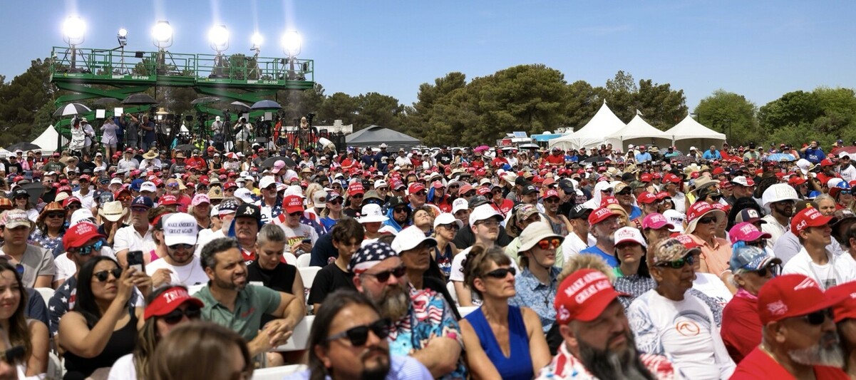 Large crowd of people wearing various hats, including many with "Make America Great Again" slogans, gathered outdoors with trees and white tents in the background, illuminated by bright lights mounted on green scaffolding.