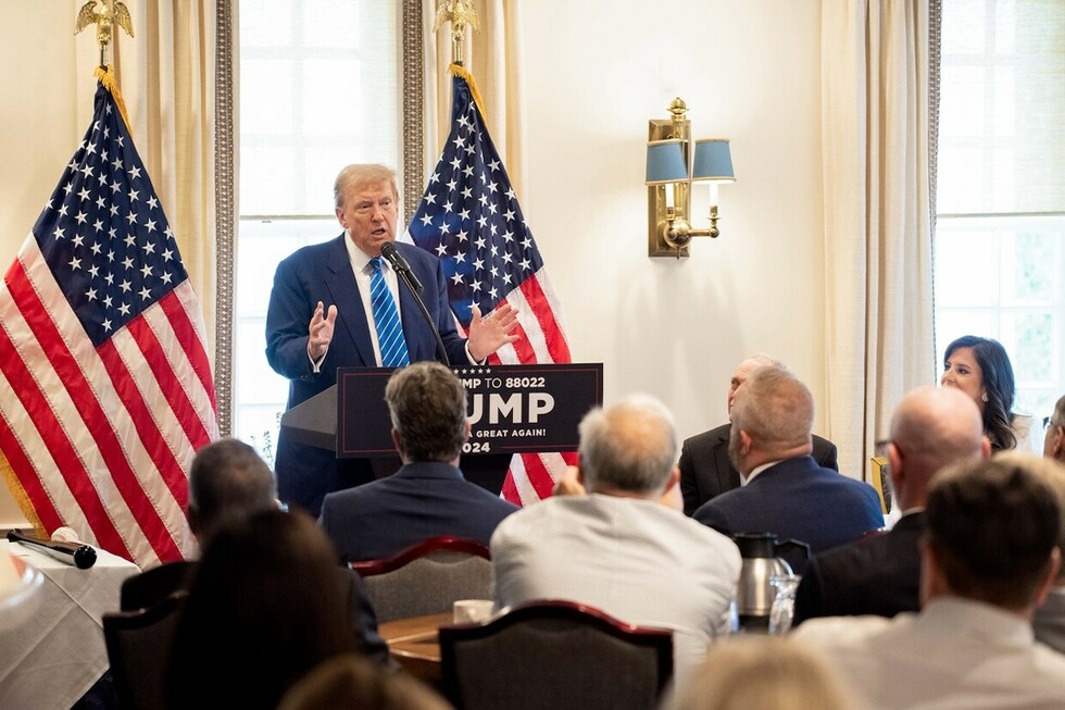 A man wearing a blue suit and blue striped tie speaks at a podium with a "TRUMP 2024" sign in front of an audience, flanked by two American flags.