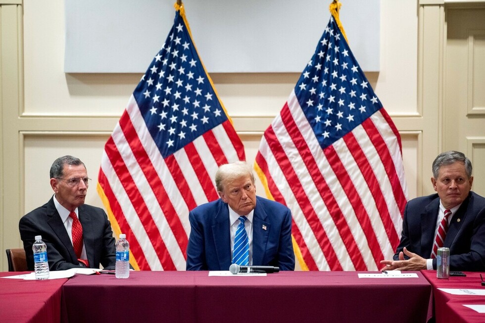 Three men sitting at a table with two American flags in the background, wearing suits and ties, and a microphone placed on the table in front of the central figure.