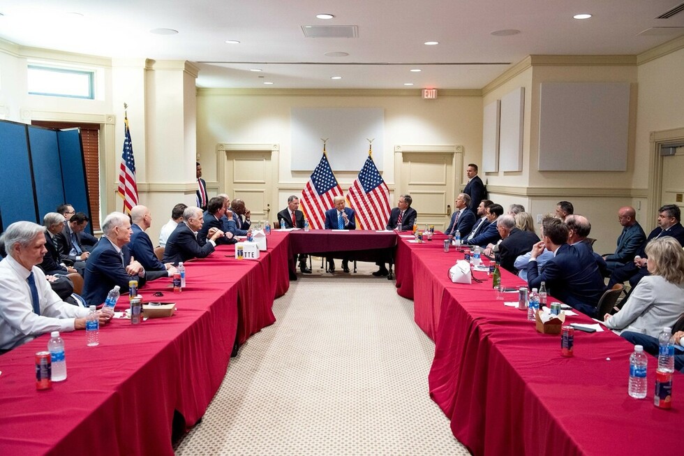A political meeting with numerous individuals seated around a U-shaped table draped in red cloths, two American flags displayed prominently at the head of the table, with three central figures engaged in discussion.