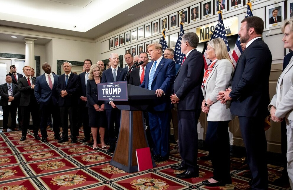 Donald Trump speaking at a podium with a sign that says, "TEXT TRUMP TO 88022, TRUMP, MAKE AMERICA GREAT AGAIN!, 2024", surrounded by a group of people, American flags, and framed photographs in the background.