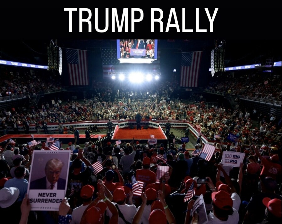 Large crowd attending a Trump rally, with supporters waving American flags and holding signs such as "Never Surrender" and "Too Big to Rig." The stage is illuminated with bright lights, and a screen above displays an image of the former president. The arena is decorated with American flags.