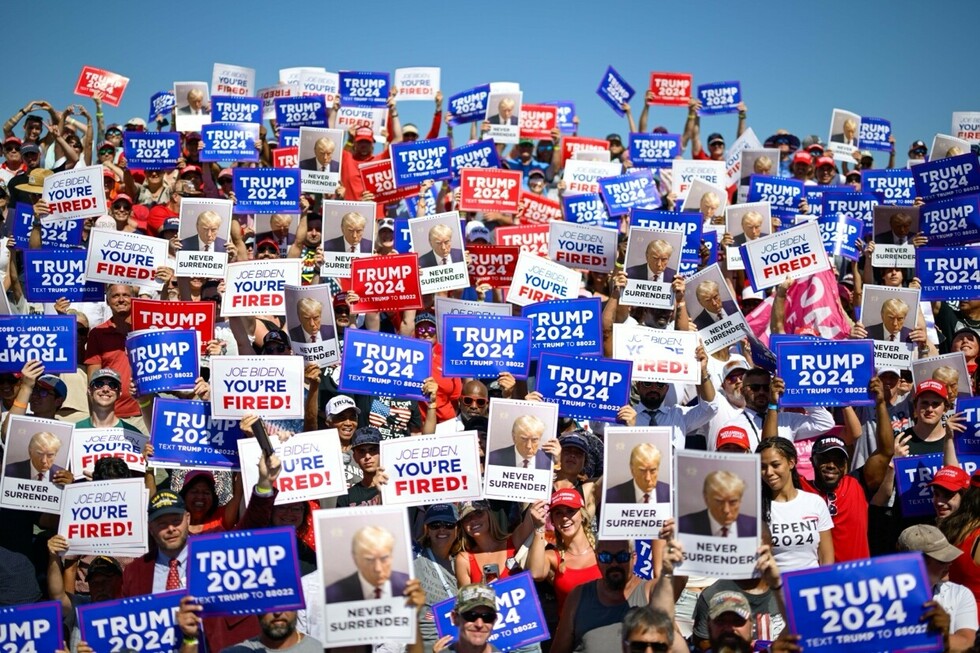A large crowd of people holding various pro-Trump signs at a rally, including messages like "Trump 2024," "Joe Biden You're Fired," and "Never Surrender," with many attendees wearing red "Make America Great Again" hats.