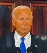 A man with white hair wearing a suit, white dress shirt, and blue tie is speaking into microphones against a wooden background.