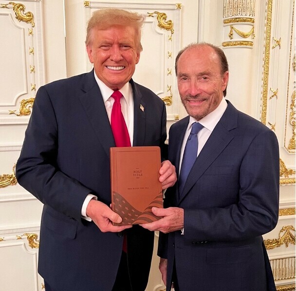 Two men in suits standing together in an ornate room, one holding a Bible with an American flag design on the cover, smiling for the camera.
