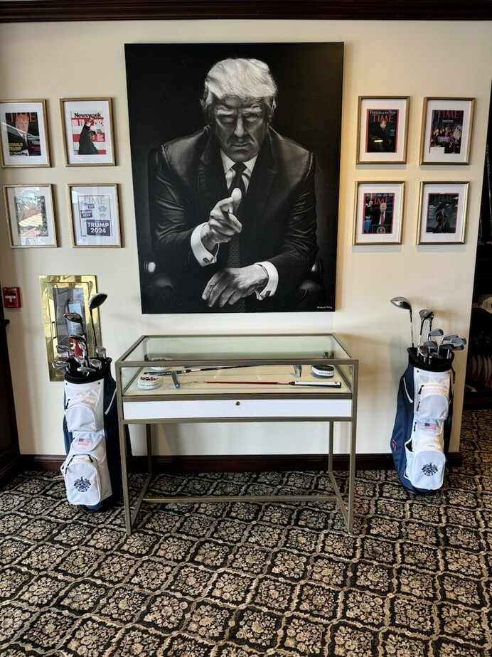 Framed black and white portrait of a man in a suit pointing forward, surrounded by framed magazine covers on the wall. Below the portrait is a glass-topped display case containing golf memorabilia, flanked by two golf bags with clubs. The room features patterned carpet and wood trim.