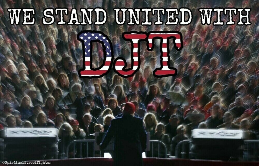 WE STAND UNITED WITH DJT; a large crowd of people, many wearing red caps, stands in support of an event, with a person addressing them from a podium in the foreground.