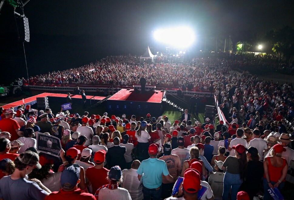 A large crowd of people wearing red hats and various apparel gather at an outdoor event with a stage. The stage is illuminated by bright lights, and a speaker stands at a podium addressing the audience. A large number of people are visible in the background, creating a densely packed scene. Some attendees are holding signs, and the event is taking place during the night.