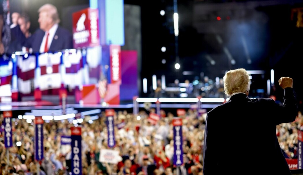 Crowd attending a political rally with a speaker on stage, large screens displaying the speaker, and banners with state names. The speaker is seen from behind in the foreground, raising a fist. The stage is adorned with American flags and vibrant lighting.