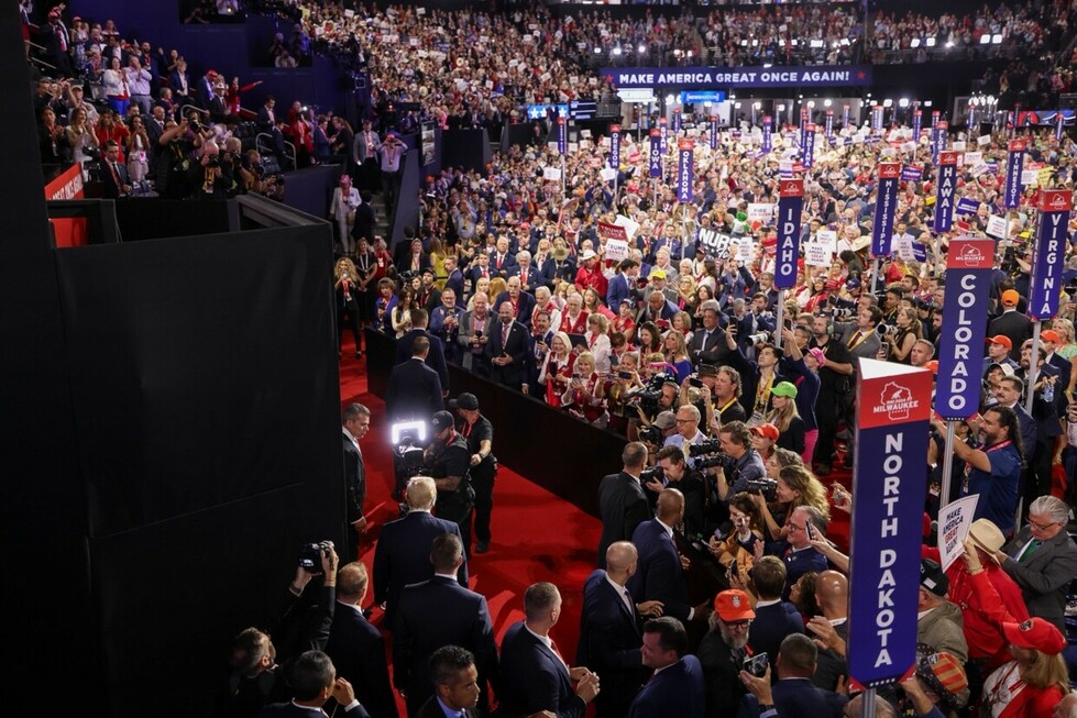 A large crowd of people attends a political rally, with prominent signs for various states like North Dakota, Colorado, and Idaho. Attendees are seen holding up signs and wearing red, white, and blue apparel. The backdrop features a "Make America Great Again" banner. Photographers and security personnel are visible in the foreground.