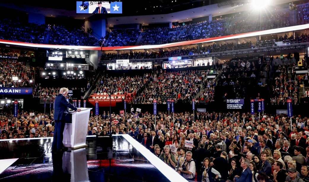 Large political rally with a speaker at the podium addressing a crowded stadium filled with supporters. Campaign signs visible, reading "Make America Great Again" and "Trump Pence." Arena lights and media coverage platforms in the background.