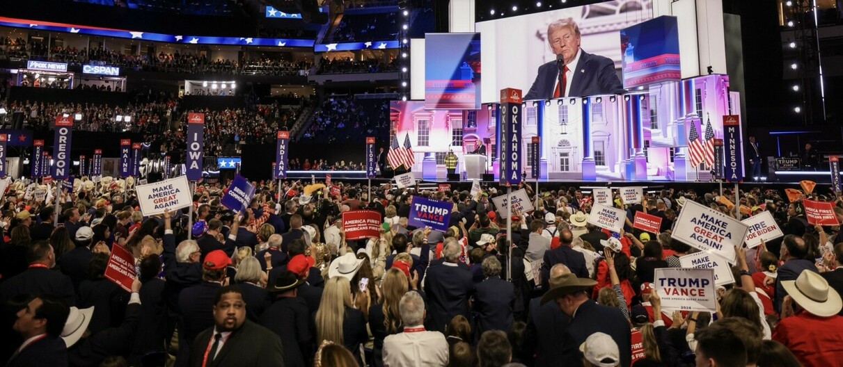 Political rally featuring a speech with a large crowd holding signs like "Make America Great Again" and "Trump Vance". Visible state markers include Oregon, Iowa, Oklahoma, Maine, Kentucky, Florida, and Minnesota. A large screen displays a speaker with an American flag backdrop. Attendees wear merchandise supporting the event and TV news channel banners are seen in the background.