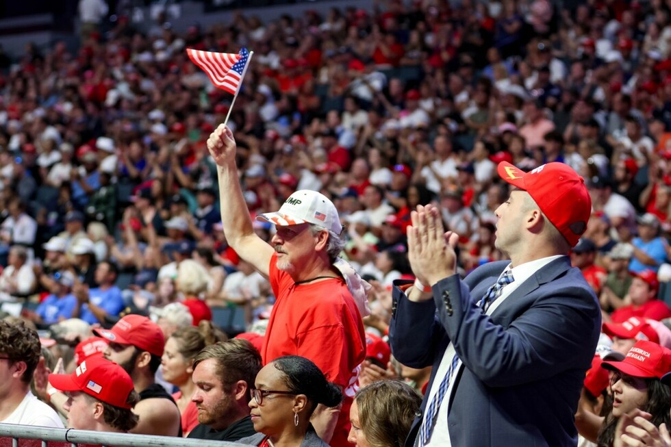 Crowd at a political rally, people wearing red hats and patriotic clothing, man holding small American flag, enthusiastic applause and cheering, large indoor venue.