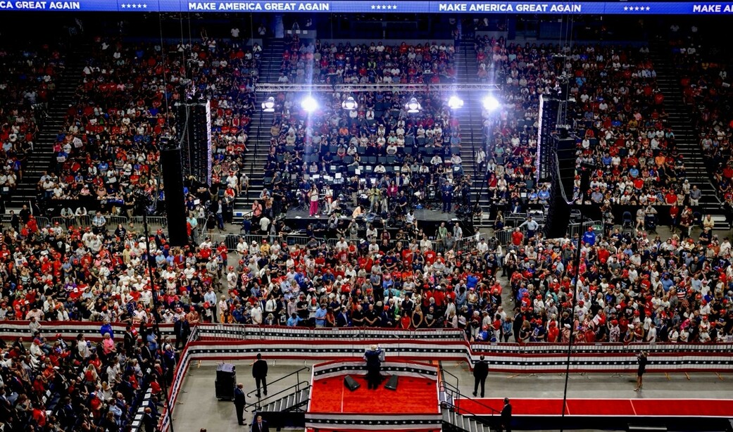 Large crowd gathered in an indoor arena, with many people wearing red shirts and hats. A stage is set up at the front of the arena, adorned with red, white, and blue decorations. A "Make America Great Again" banner is prominently displayed at the top of the image. Stadium lights illuminate the scene, and there are various barriers and cameras set up, indicating a significant public event or rally.
