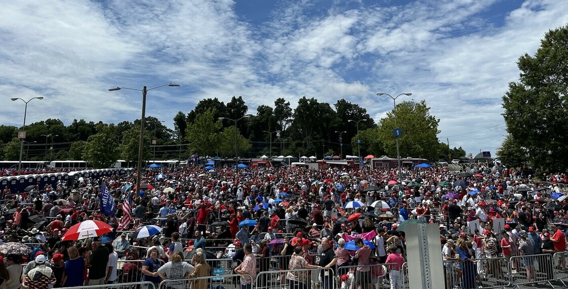 Large crowd gathered outdoors under a partly cloudy sky, with people holding umbrellas and wearing red caps, surrounded by trees and portable toilets.