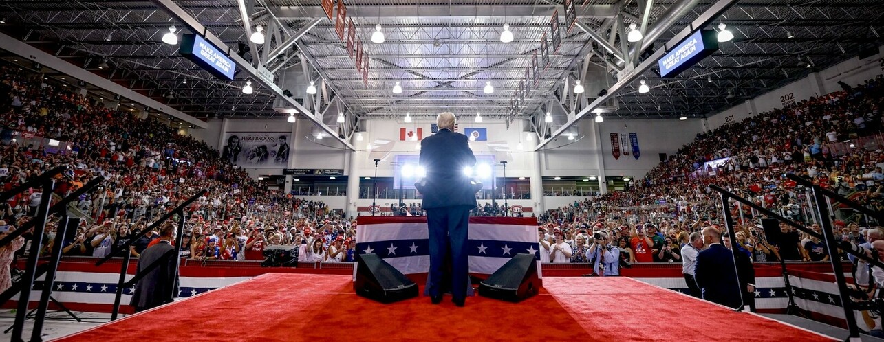 Large indoor rally with a speaker standing on a stage decorated with American flags, facing a cheering crowd. The audience fills the entire arena, with some people holding signs and taking photos. Overhead signage reads "MAKE AMERICA GREAT AGAIN," and the rafters are adorned with flags and banners. Bright lights illuminate the stage, highlighting the speaker's presence.