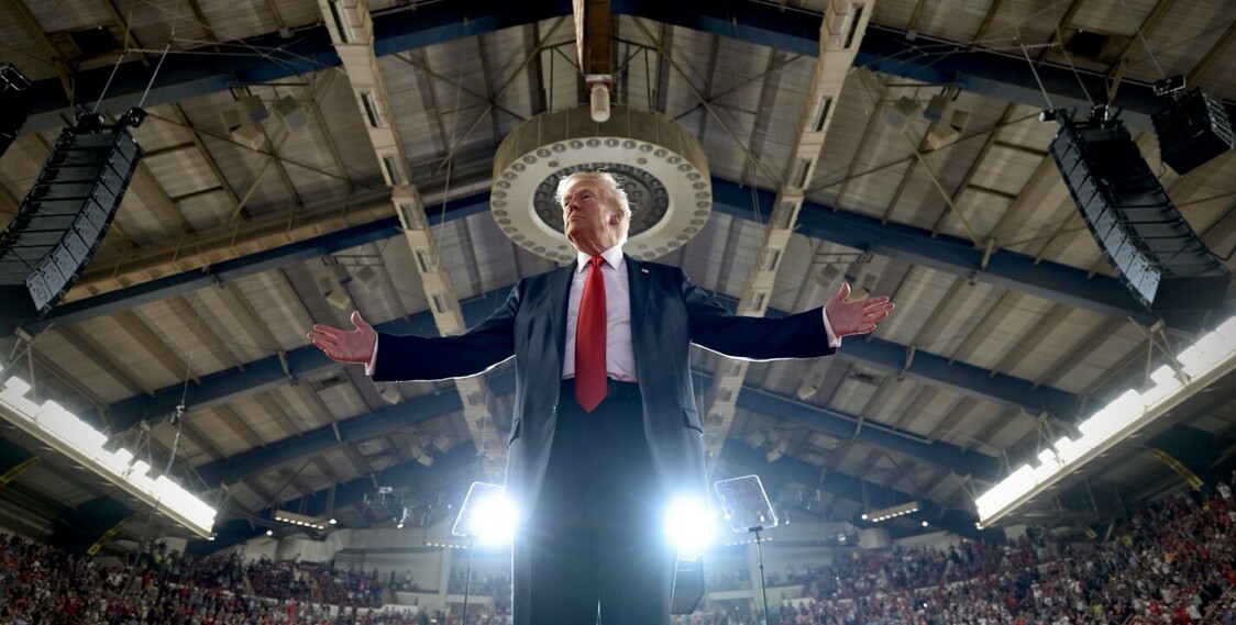 Man in a suit with a red tie stands on a stage with arms outstretched during a large indoor event, under a high, arched ceiling with exposed beams and lighting fixtures; audience visible in the background.