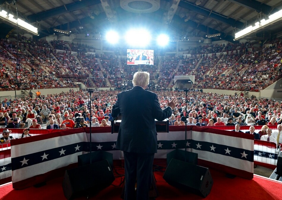 A speaker addresses a large and packed audience in an indoor stadium, standing on a stage adorned with patriotic-themed banners and flags. Bright lights illuminate the scene from above, and there are teleprompters on both sides of the podium. The audience consists of a diverse group of people, many wearing red attire or accessories, and it appears engaged and attentive. A large screen is visible in the background displaying a live feed of the event.