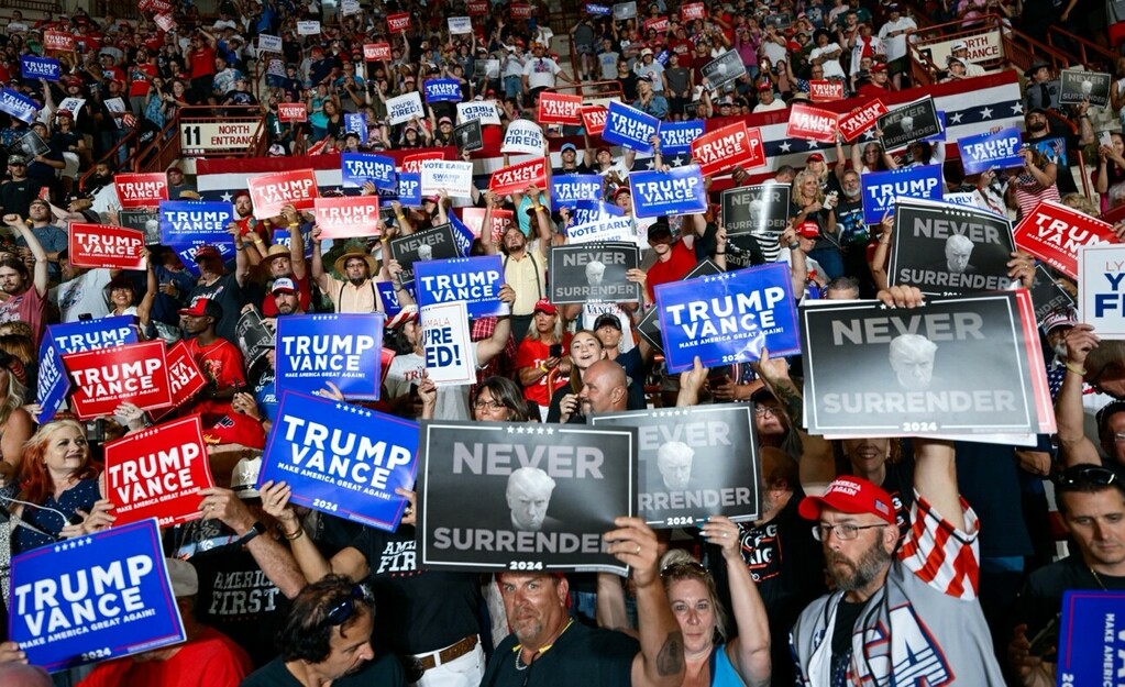 A large crowd of people holding signs that read "Trump Vance 2024," "Never Surrender," "Make America Great Again," "You're Fired!" and "Vote Early" at a political rally. The crowd is energetic, with many individuals wearing red hats and patriotic clothing, creating a vibrant and packed atmosphere indoors.