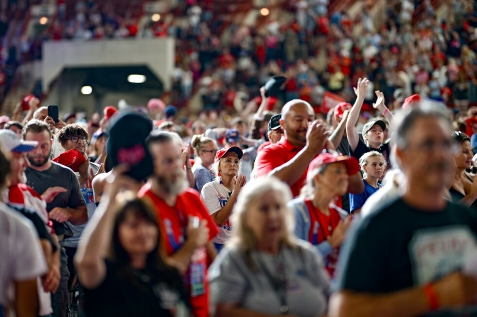Large crowd of people at an indoor event, many wearing red shirts and hats, some clapping and capturing moments with their phones, with a focus on an older woman in a white shirt clapping.