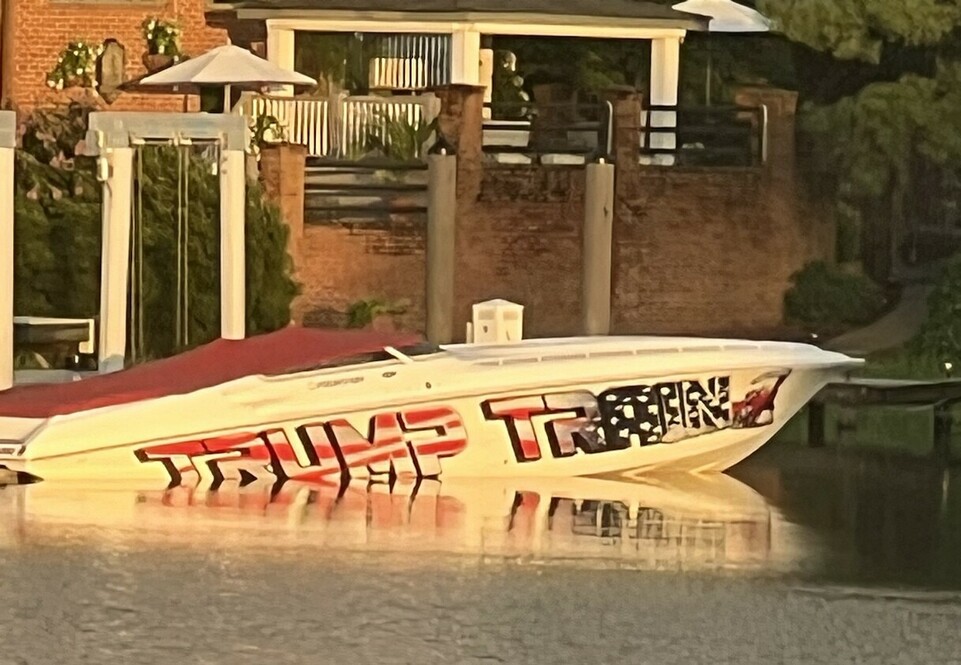 Speedboat with the phrase "Trump Train" in large, bold letters anchored near a brick house with a covered porch and a white umbrella on the dock.