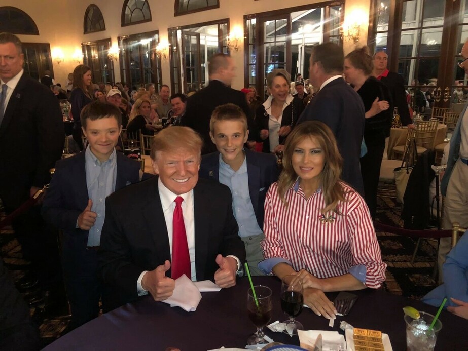 A man in a dark suit with a red tie and a woman in a striped shirt are seated at a table in a crowded indoor event, smiling at the camera. Two young boys are standing beside them, both giving thumbs-up gestures. The background shows people seated at tables, engaged in conversations, and a man standing guard. The atmosphere appears lively and festive.