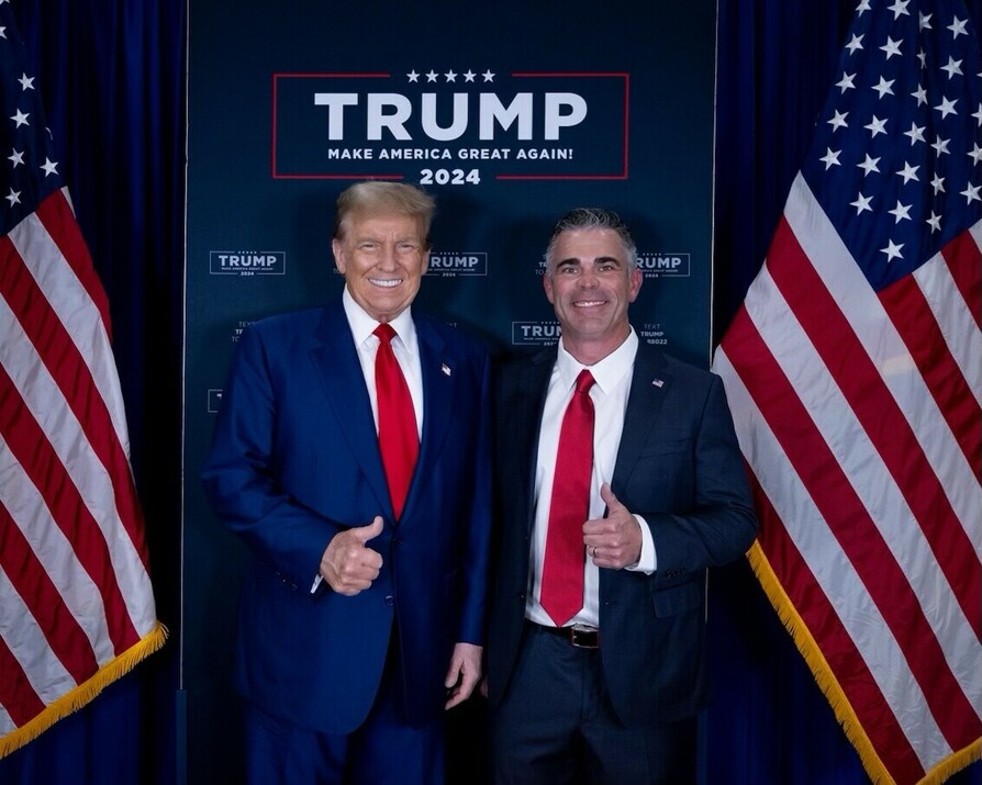 Two men wearing suits, standing side by side, both giving thumbs-up gestures. They are positioned in front of a "Trump 2024" campaign banner with the slogan "Make America Great Again!" Two American flags are placed symmetrically on either side of the banner.