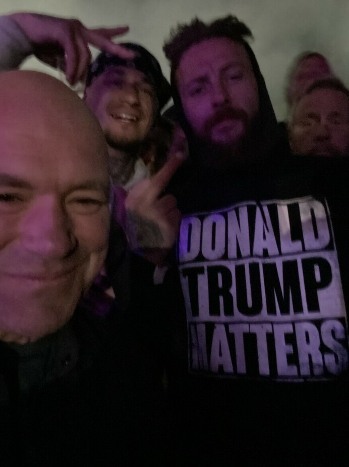 A group of people posing for a selfie, with one person wearing a hoodie that has "DONALD TRUMP MATTERS" printed on it. The background is dark, and the group appears to be in a casual or social setting. One person is making a peace sign gesture.