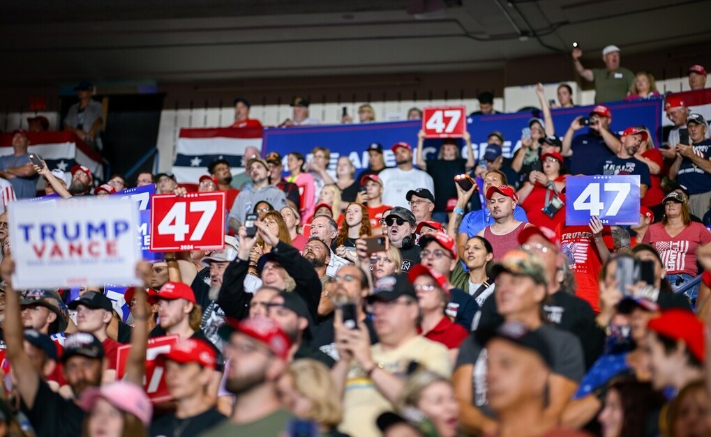 A large crowd of people, many wearing red hats, holding signs that read "47" and "TRUMP VANCE," gathered in an indoor venue adorned with red, white, and blue decorations.
