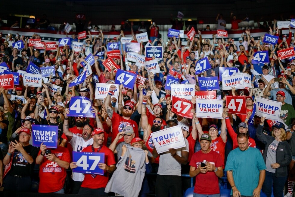 A large crowd of enthusiastic supporters holding signs with slogans such as "47," "Trump Vance," and "Make America Great Again" at a political rally. Many attendees are wearing red hats and shirts, with hands raised in support.