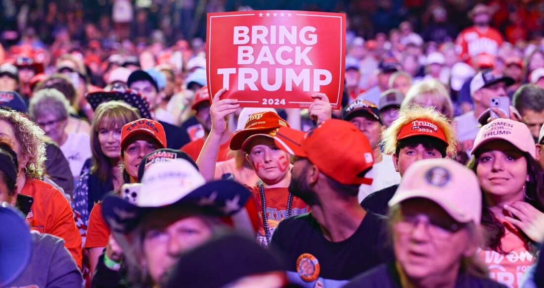 Crowd of people at a political rally, with central figure holding a "Bring Back Trump 2024" sign, many wearing red Trump hats and shirts.