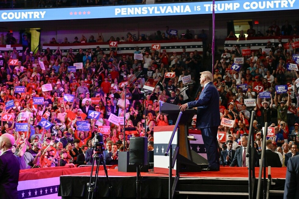 Man speaking at a podium during a large political rally with a crowd holding various signs including "TRUMP MAKE AMERICA GREAT AGAIN," "47," "FIGHT FIGHT FIGHT," "NO MORE BULLIES," and others. The venue features a banner reading "PENNSYLVANIA IS TRUMP COUNTRY" and American flag decorations.