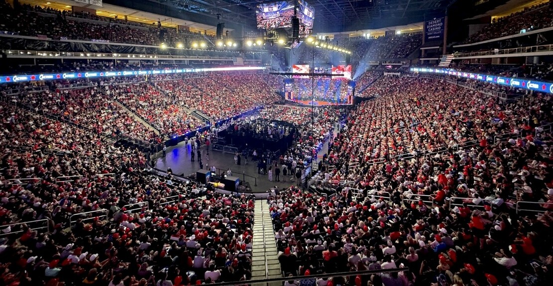 A packed sports arena filled with thousands of people, centered around a well-lit stage with LED screens displaying vibrant visuals. The crowd appears to be highly engaged, creating a dynamic and energetic atmosphere in the large indoor venue.