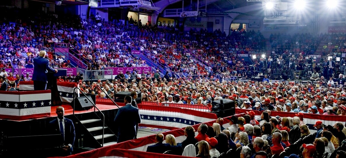 A large indoor arena filled with a crowd attending a political rally. The stage is decorated with flag-themed bunting, and the speaker is addressing the audience. Many attendees are wearing red caps, and media personnel with cameras are visible in the background. Bright lights illuminate the stage, and security personnel are present around the area.