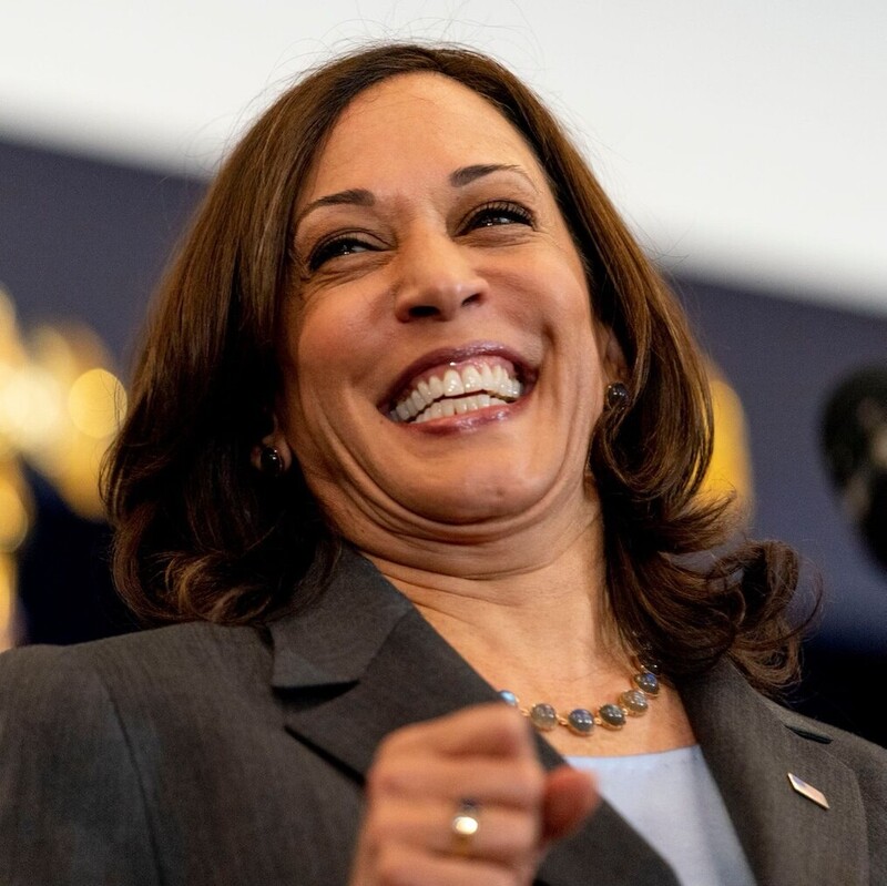 Woman with shoulder-length brown hair smiling widely during a speech, wearing a gray blazer and a beaded necklace.